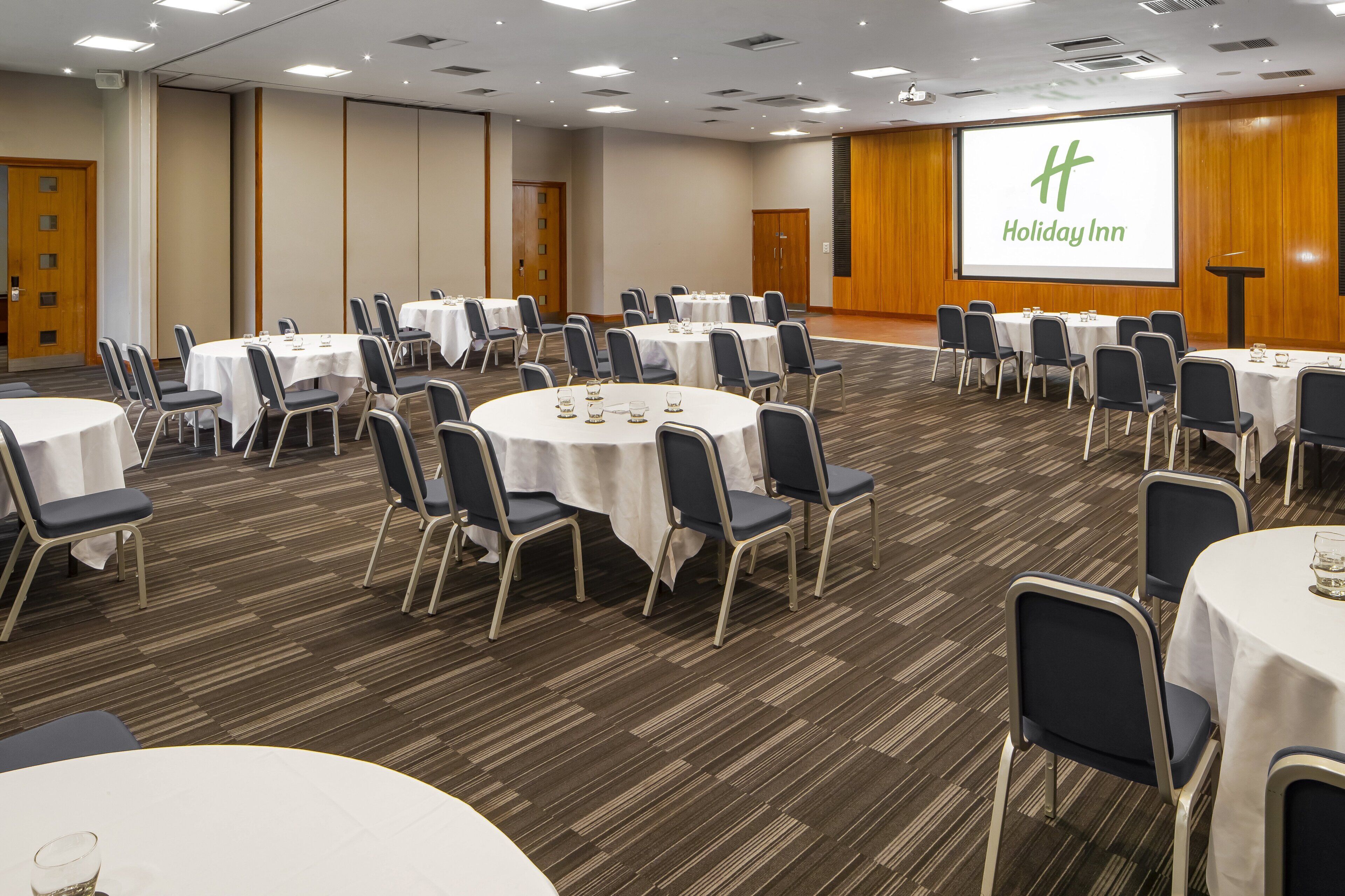 A conference room with multiple round tables and chairs, a large screen displaying the Holiday Inn logo, and a podium.