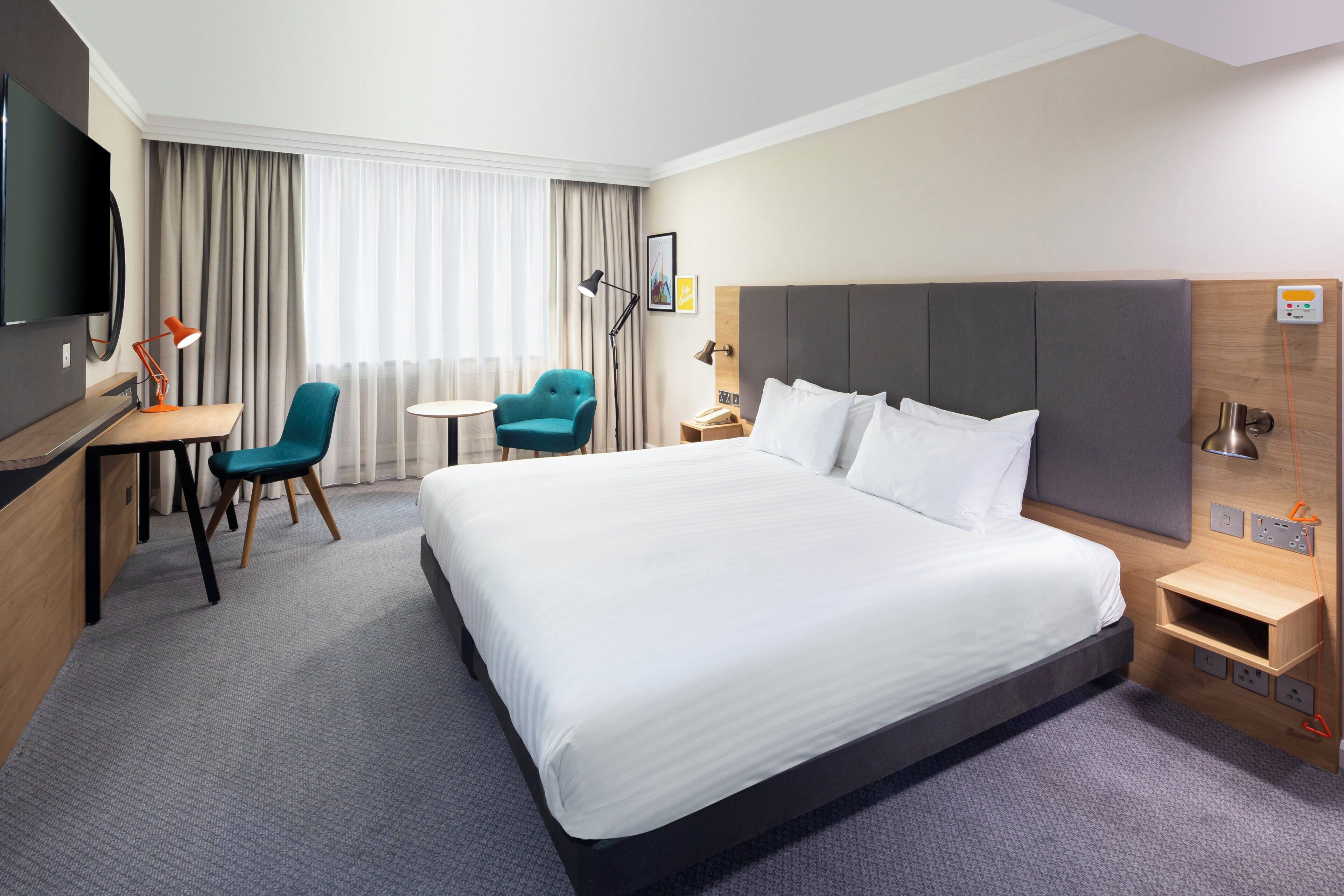 A hotel room with a large bed, a desk, a chair, a TV, and a window with curtains.