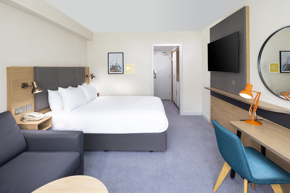 A hotel room with a bed, a desk, a chair, a TV, and a bedside table with a lamp.