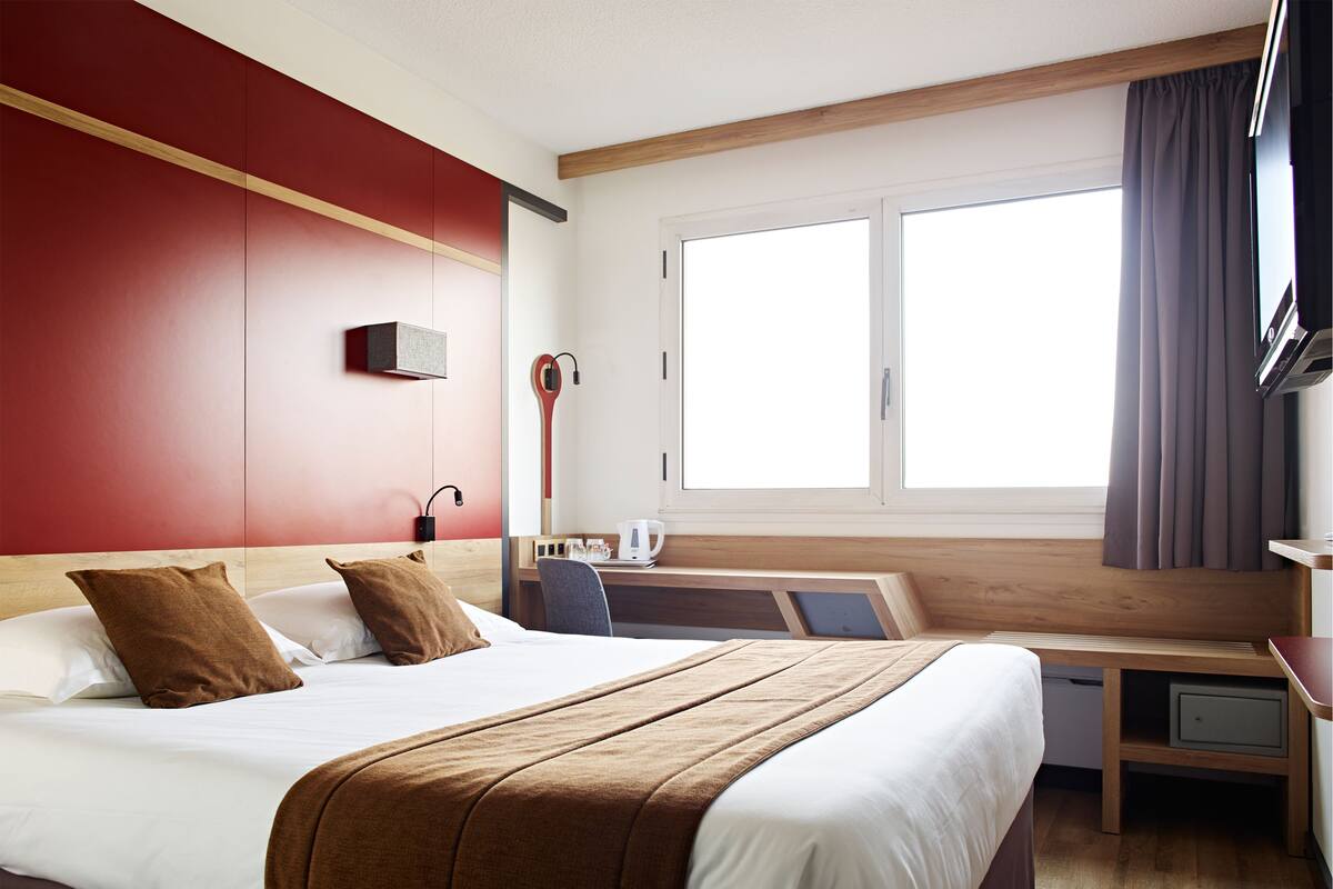 A hotel room with a bed, a desk, a chair, a TV, and a window with curtains.