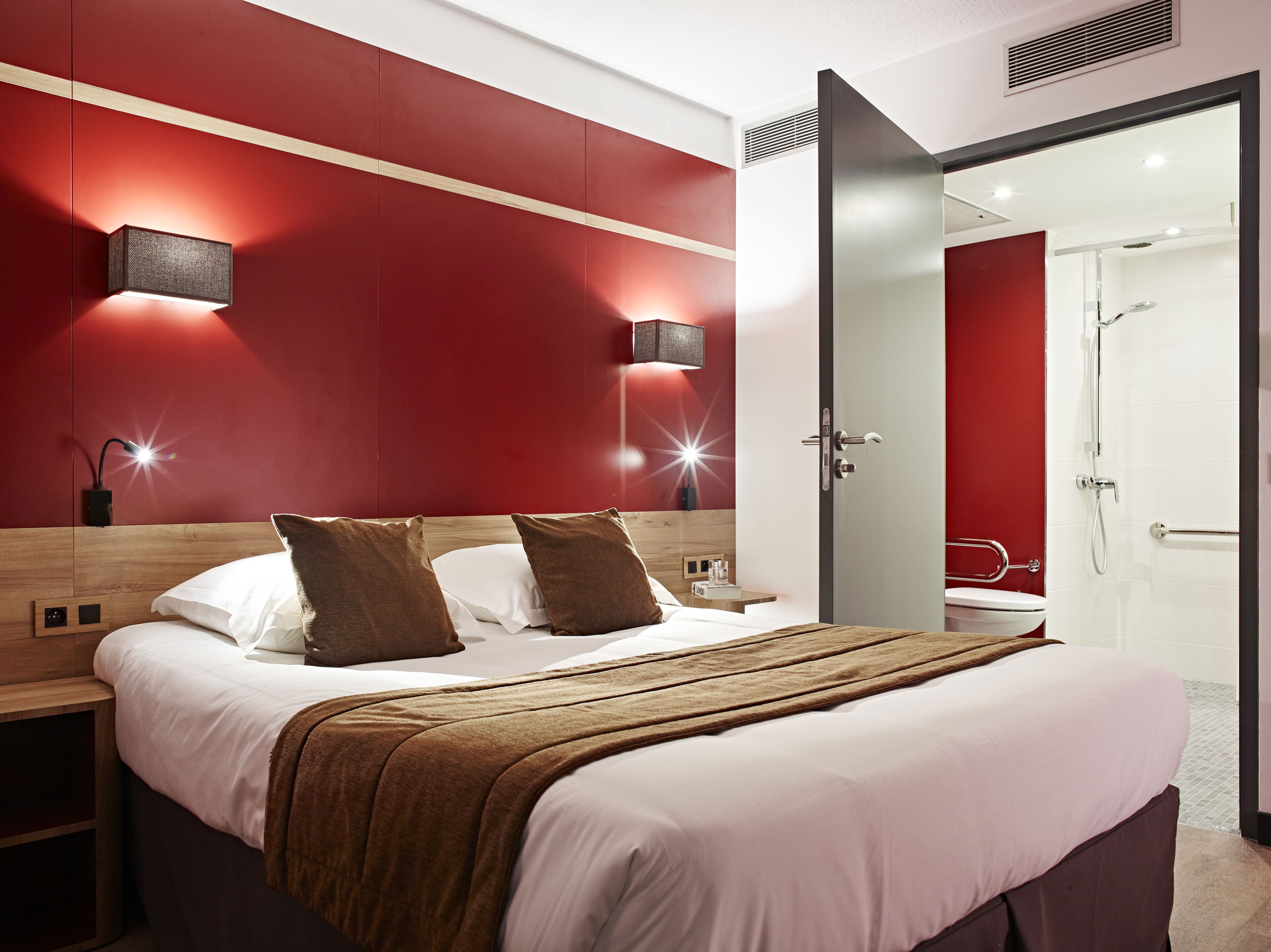 A modern hotel room with a large bed, two bedside tables, a nightstand, a red accent wall, and a bathroom visible through an open door.