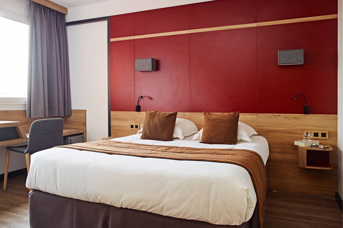 A hotel room with a large bed, a desk, and a chair. There are two bedside tables with lamps and a small shelf with glasses and a water bottle.
