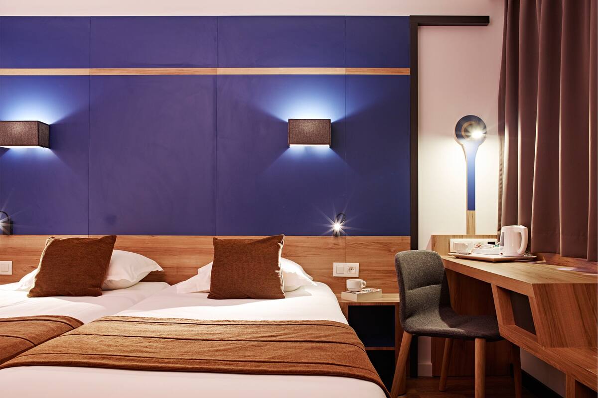 A hotel room with a bed, bedside table, lamp, chair, and a desk with a kettle.