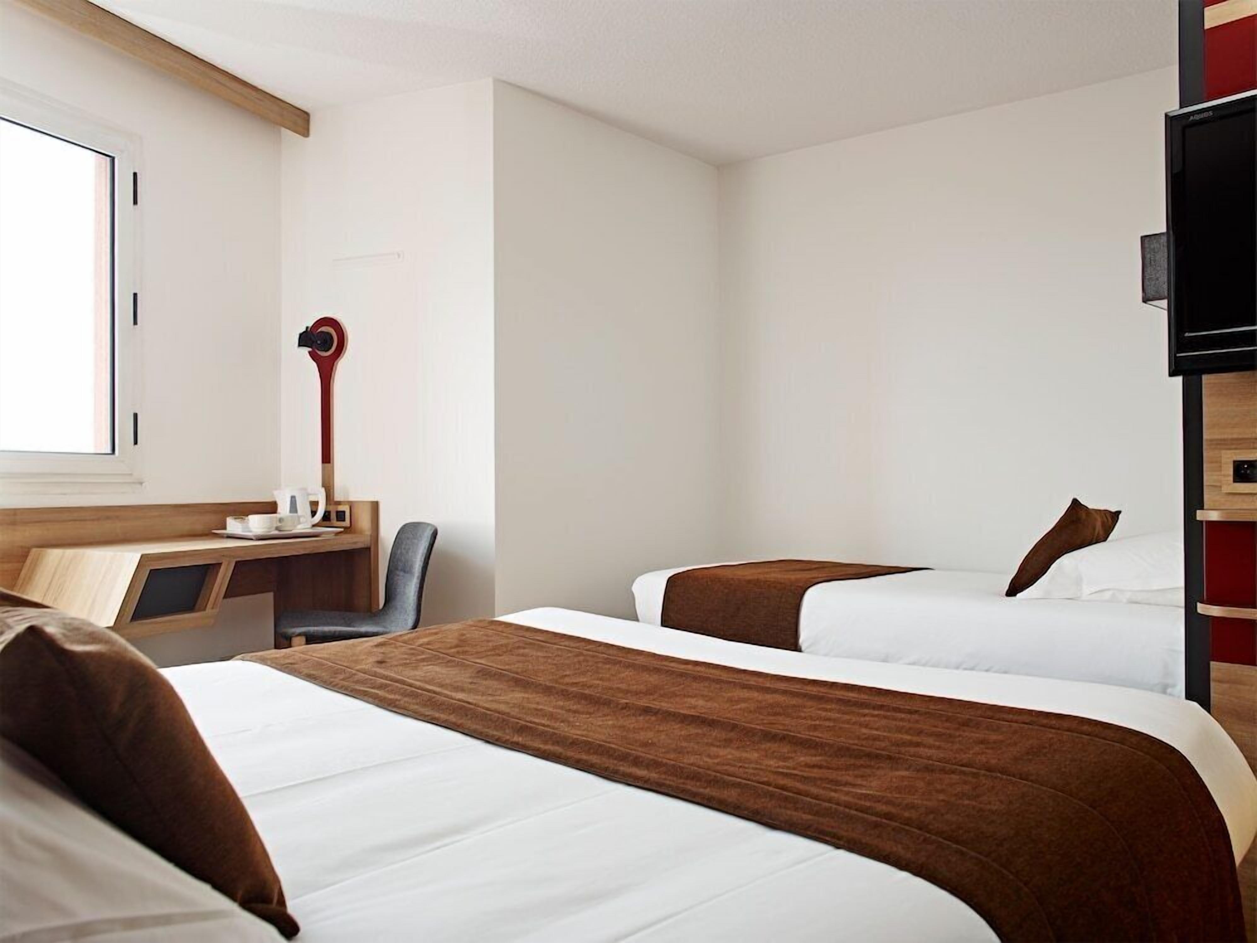 A hotel room with two beds, a desk with a chair, a TV, and a window.