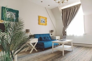 Un salon moderne avec un canapé bleu, une table basse blanche et un ordinateur portable posé sur la table. On y trouve également une plante, un tableau encadré au mur et une fenêtre avec des rideaux.