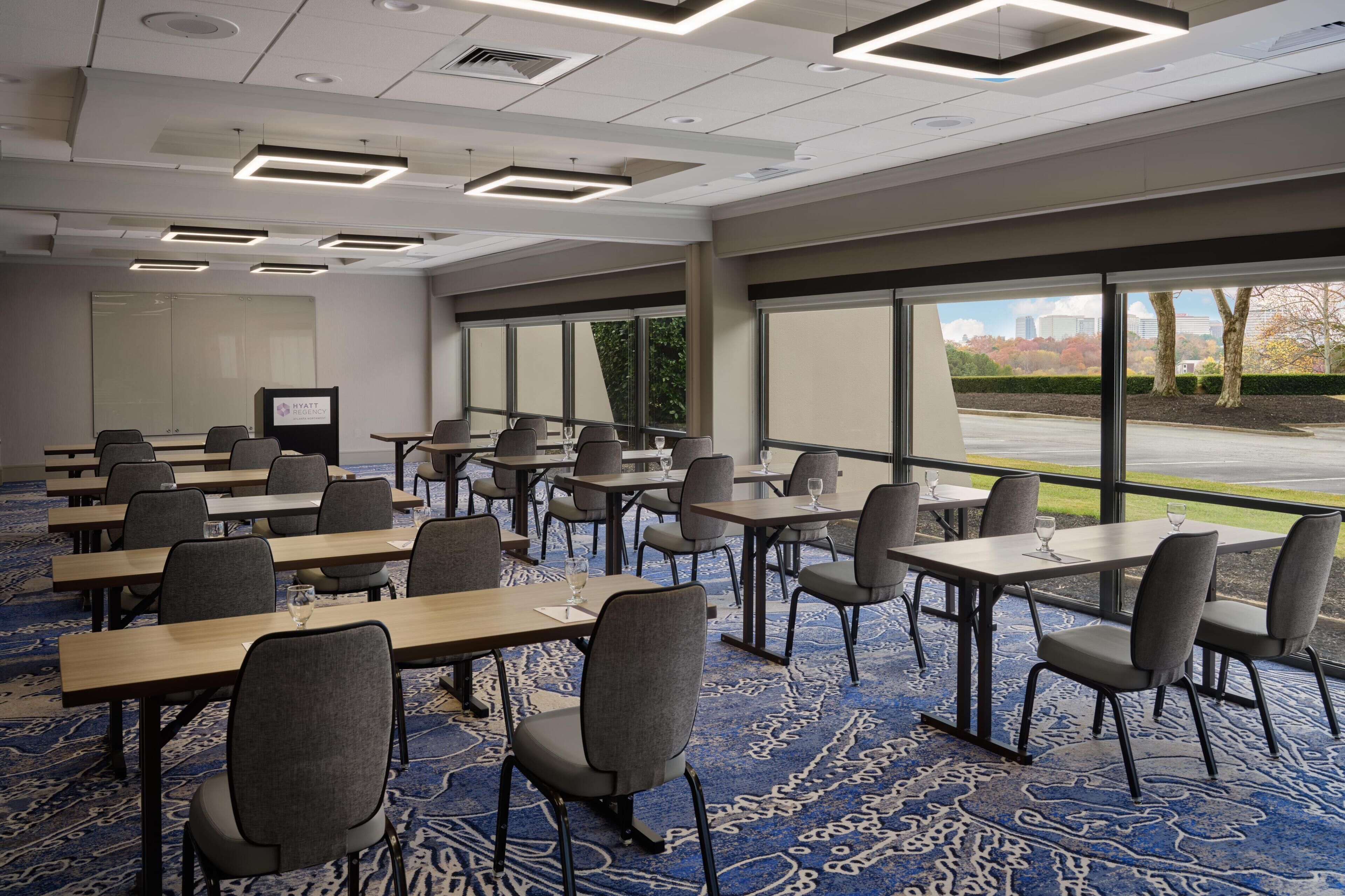A conference room with multiple tables and chairs, large windows, and a carpeted floor.
