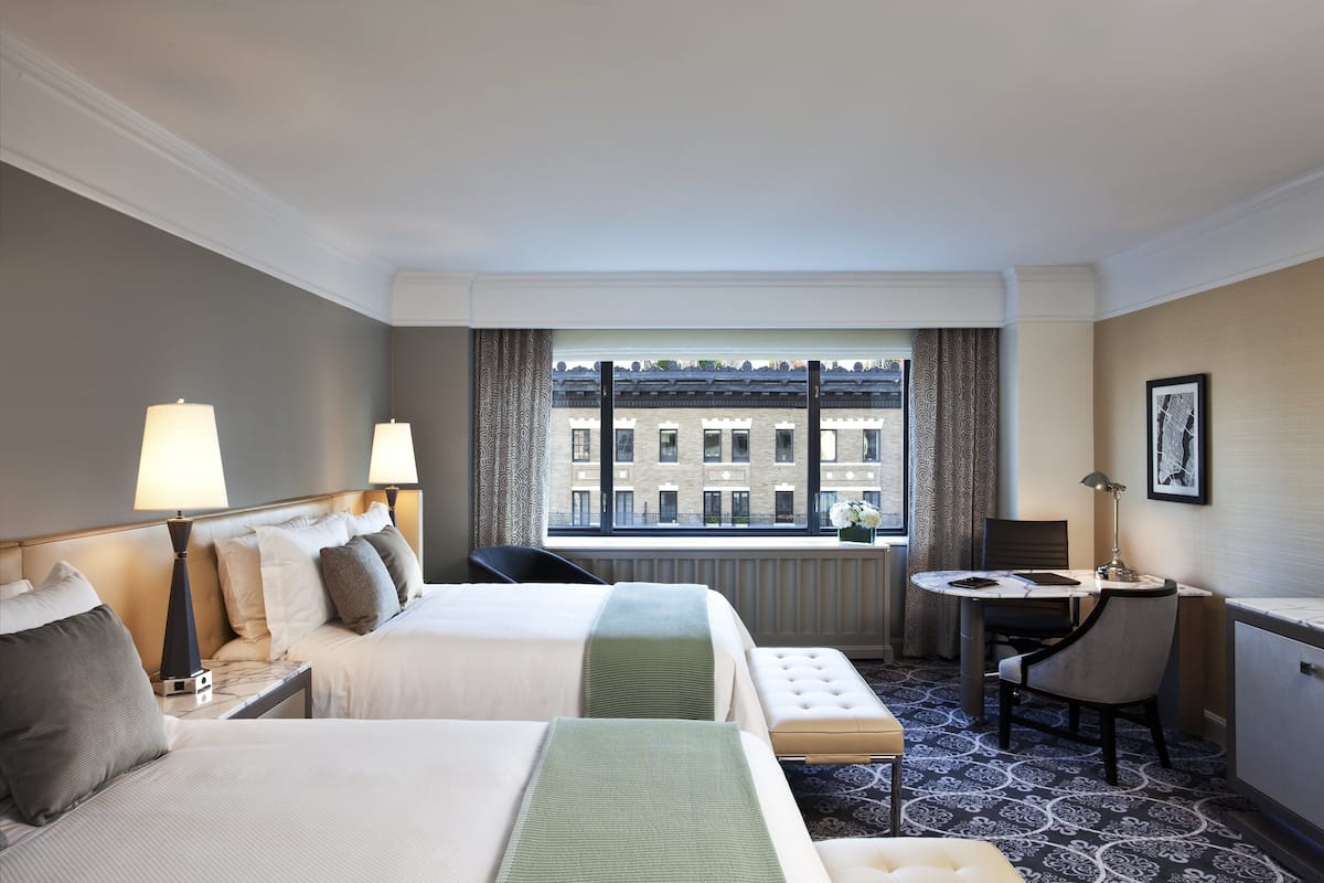 A hotel room with two beds, a dining table, a chair, and a view of a building through the window.