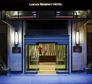 Exterior do Hotel Loews Regency, com entrada moderna destacada por portas de vidro e uma decoração floral vermelha.