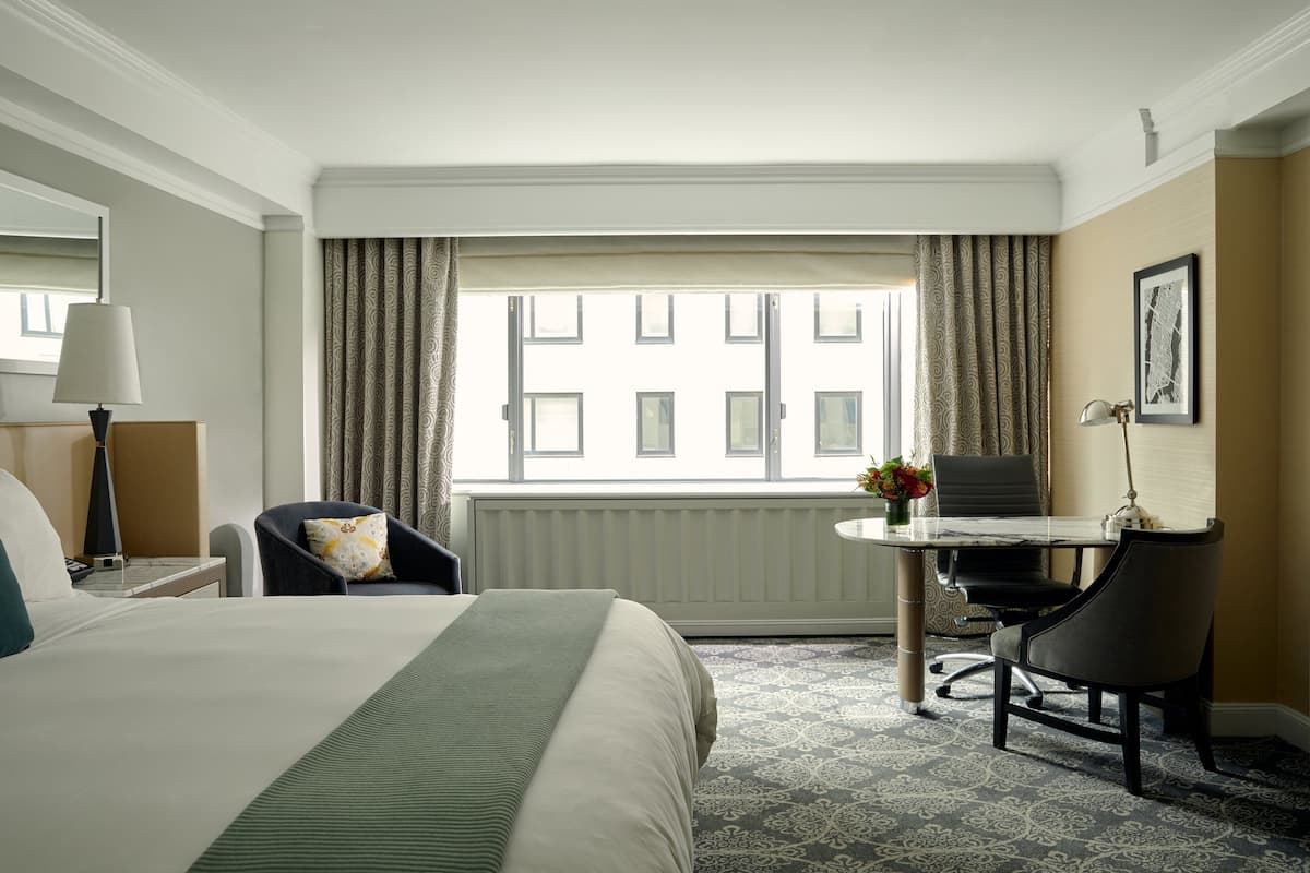 A hotel room with a bed, a desk with a chair, a lamp, a window with curtains, and a radiator.