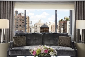 Uma sala de estar moderna com um sofá cinzento, almofadas estampadas e um arranjo floral sobre uma mesa de vidro. Uma janela grande oferece vista para edifícios da cidade.