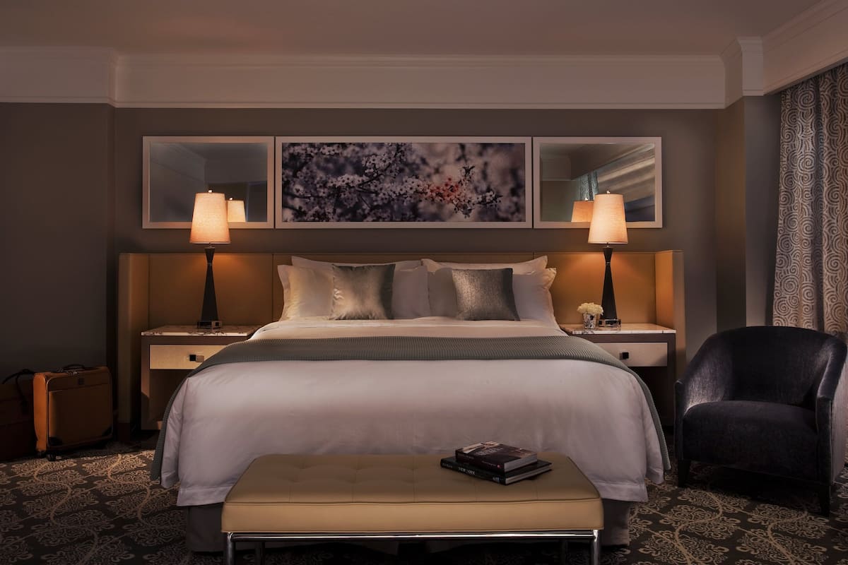 A hotel room with a large bed, two bedside lamps, a nightstand, a chair, a small table with a book, and a decorative wall piece.