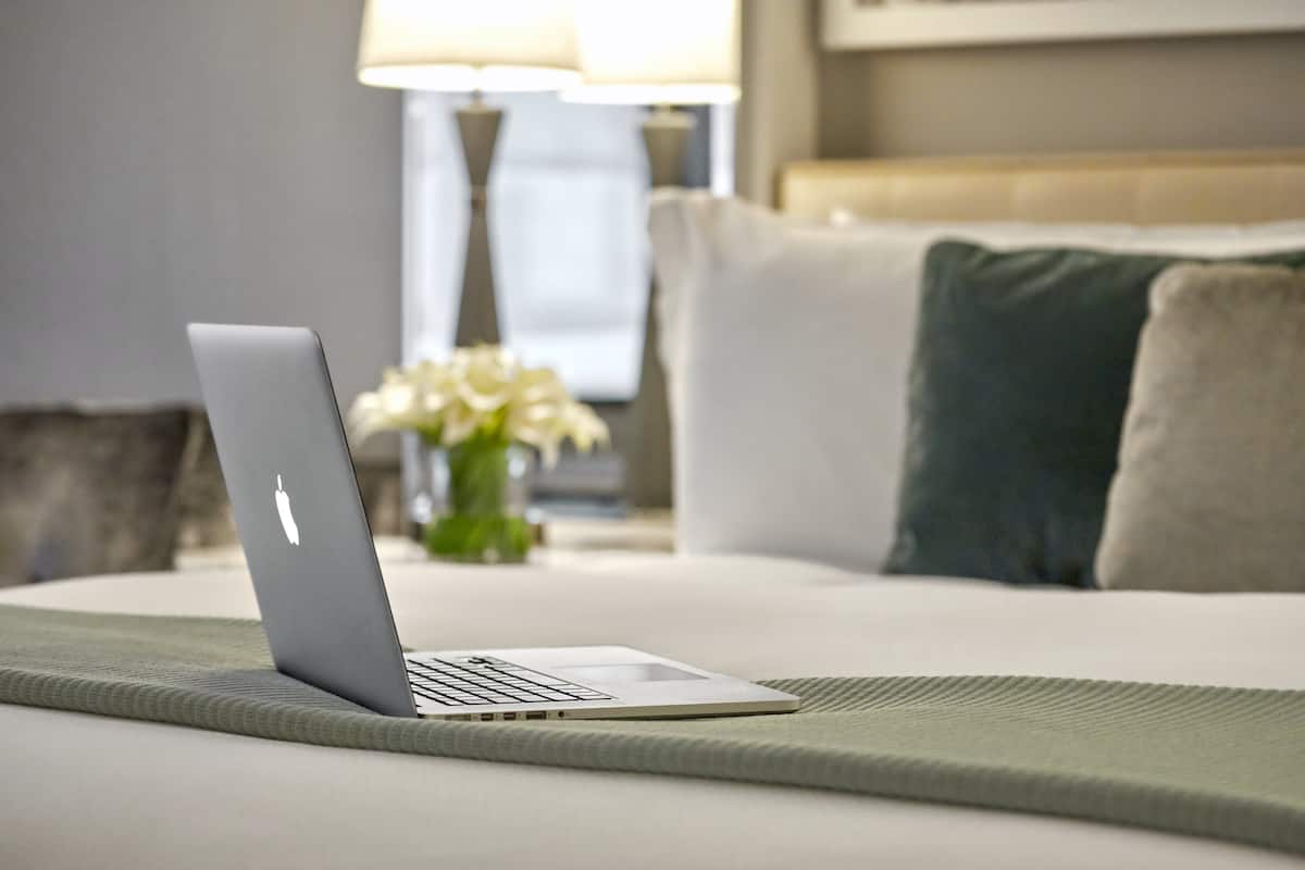 A laptop on a bed with a green bedspread, white pillows, and a lamp in the background.