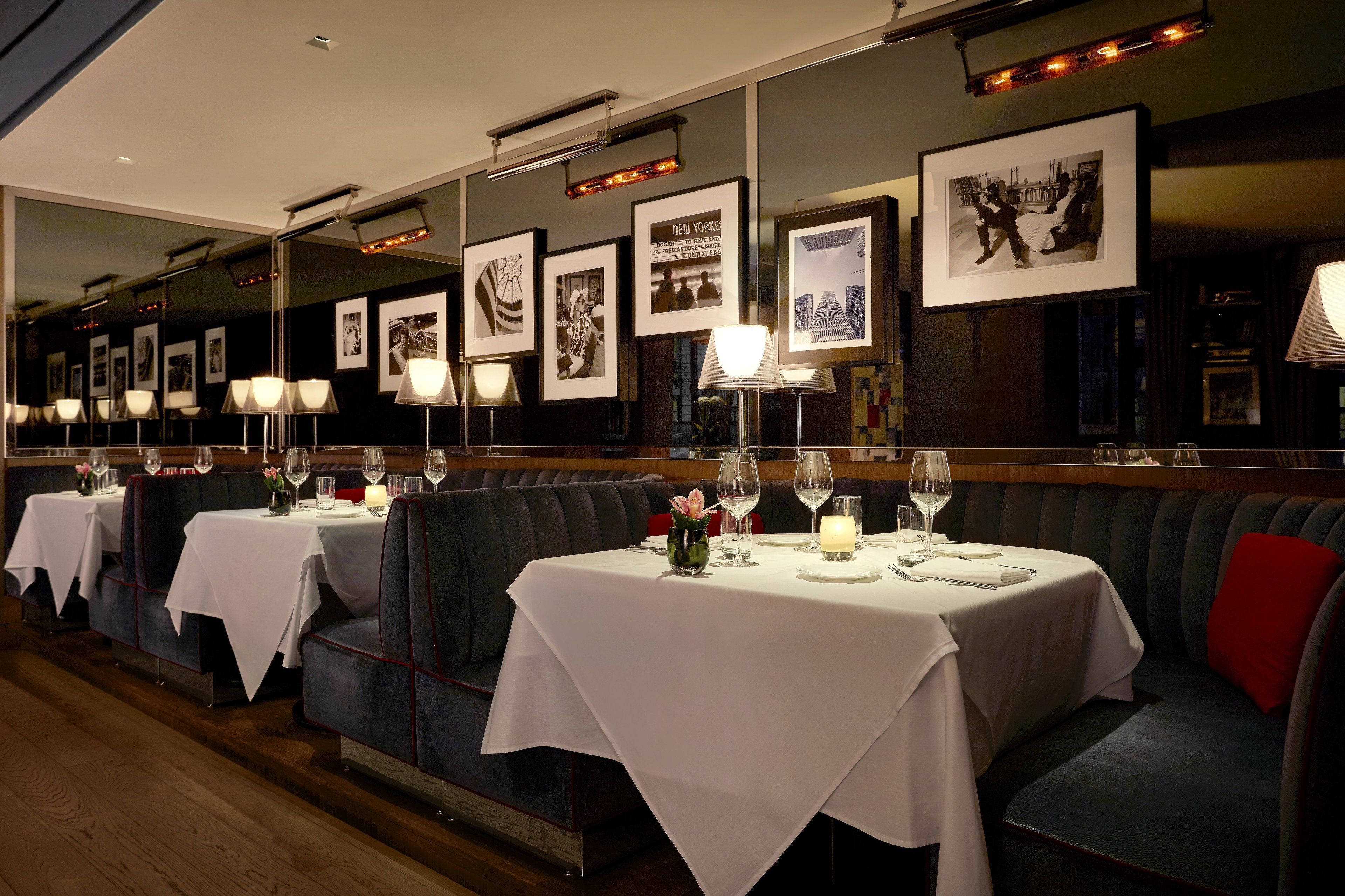 A restaurant with multiple tables, each set with white tablecloths, wine glasses, and candles. The walls are adorned with framed pictures.