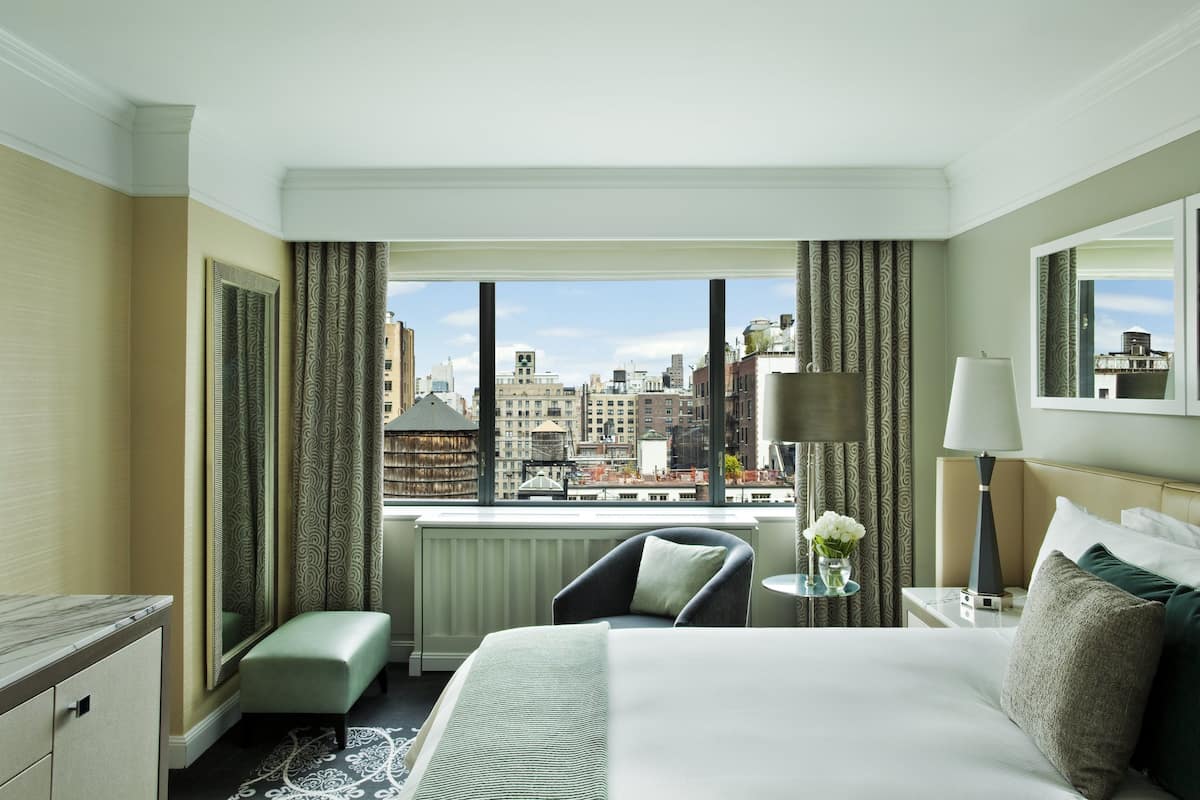 A hotel room with a large window, a bed, a chair, a nightstand, and a small table with a vase of flowers.