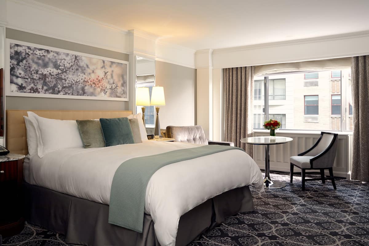 A hotel room with a large bed, a small round table, a chair, and a window with curtains.