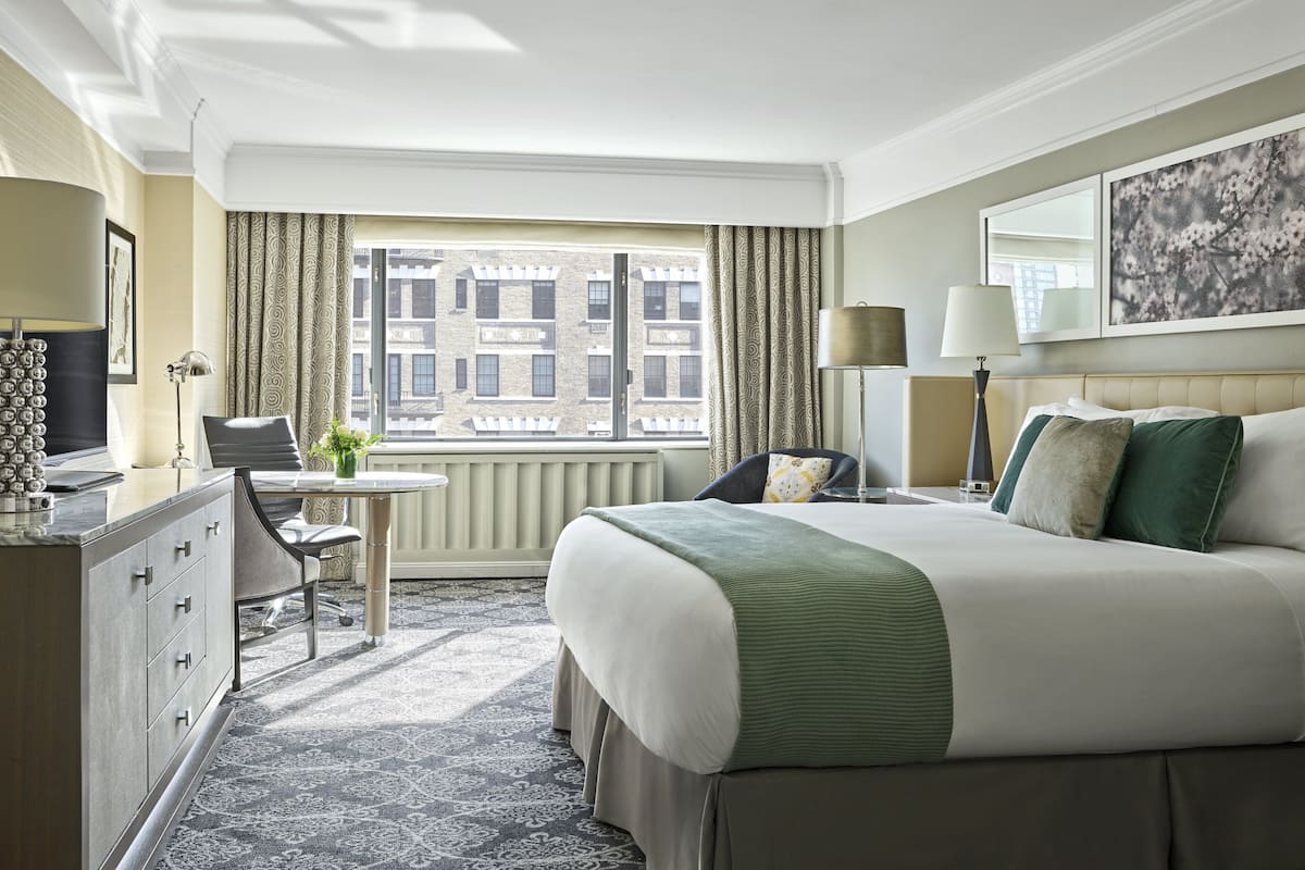 A hotel room with a large bed, a desk with a chair, a small table with a plant, a radiator, and a window with curtains.