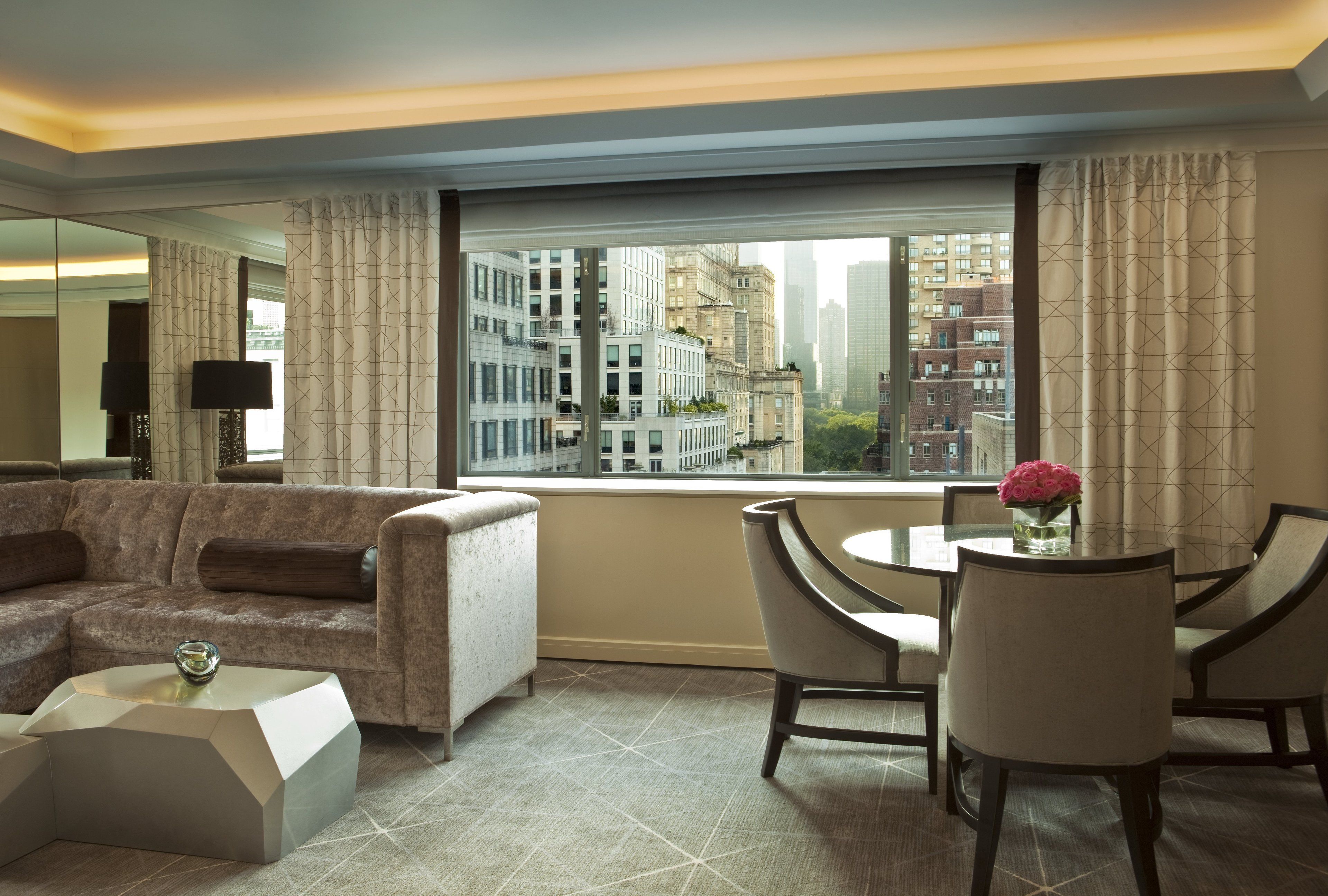 A modern hotel room with a large window, a sofa, a dining table with chairs, and a small side table.