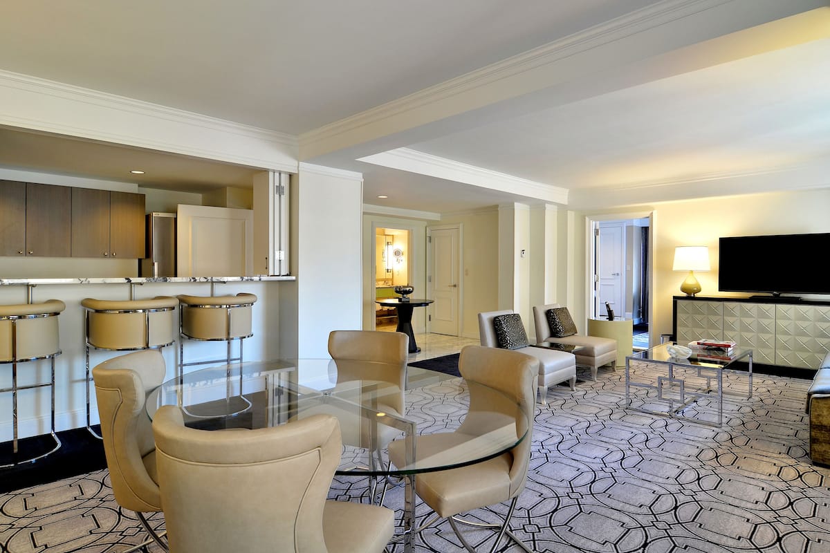 A modern hotel lobby with a glass table, leather chairs, a sofa, a TV, and a patterned carpet.