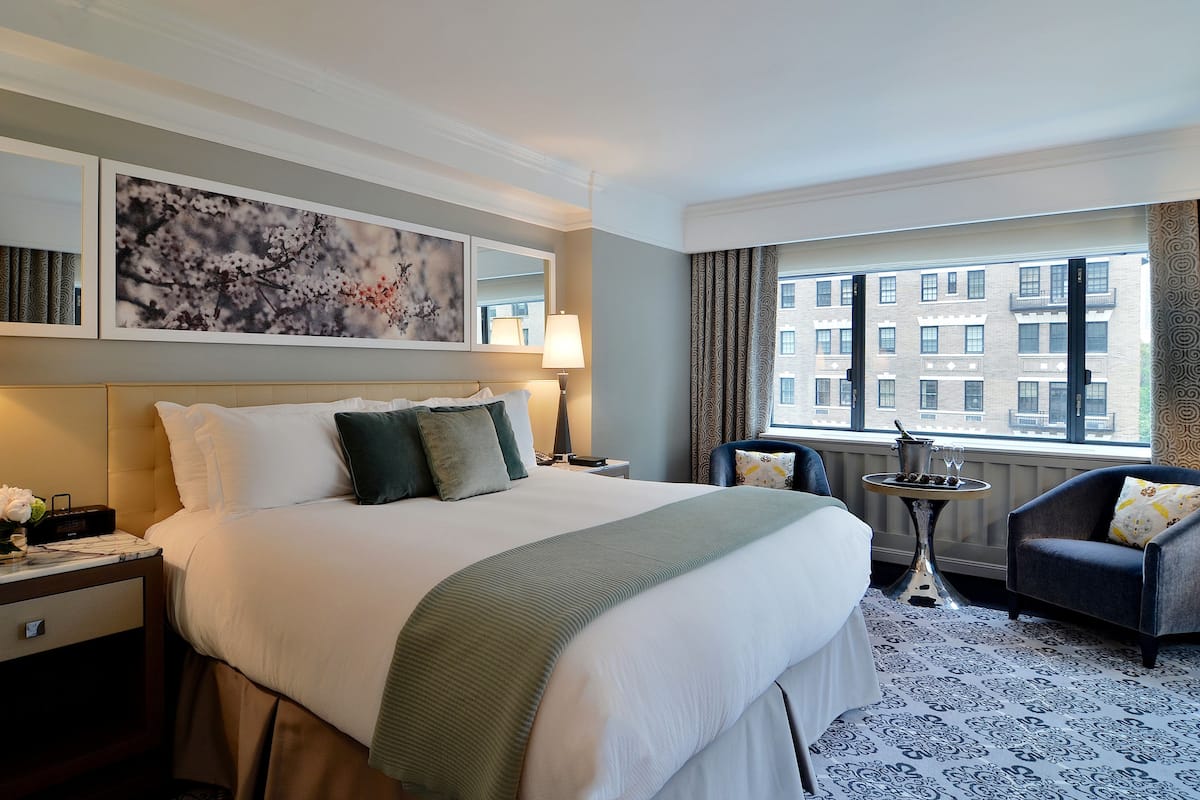 A hotel room with a large bed, two bedside lamps, a small table with a bottle and glasses, a chair, and a window with a city view.