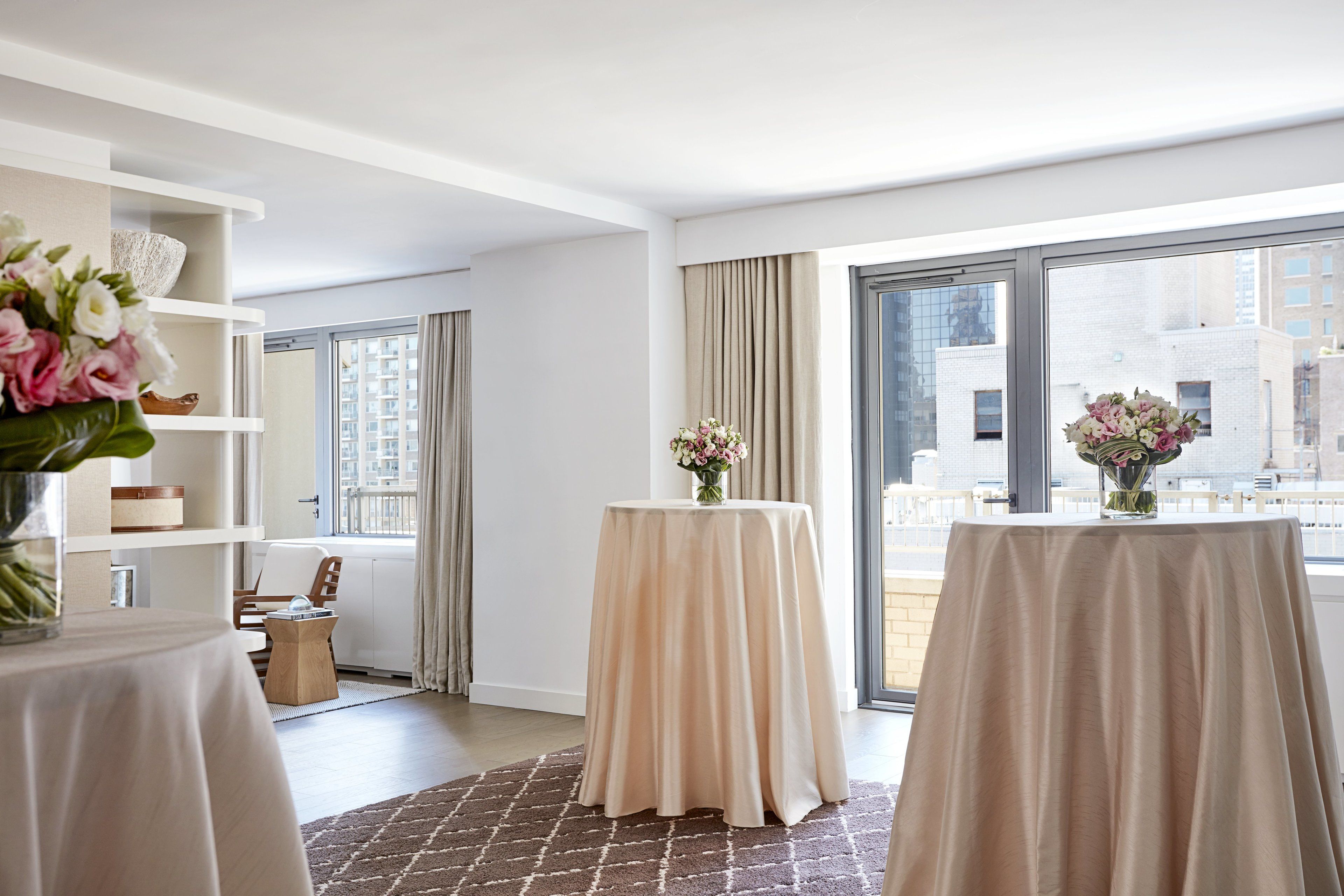 A modern hotel room with a large window, a round table with a beige tablecloth, and a bookshelf with decorative items.