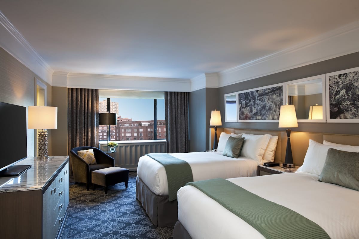 A hotel room with two beds, a desk, a chair, and a large window with a city view.