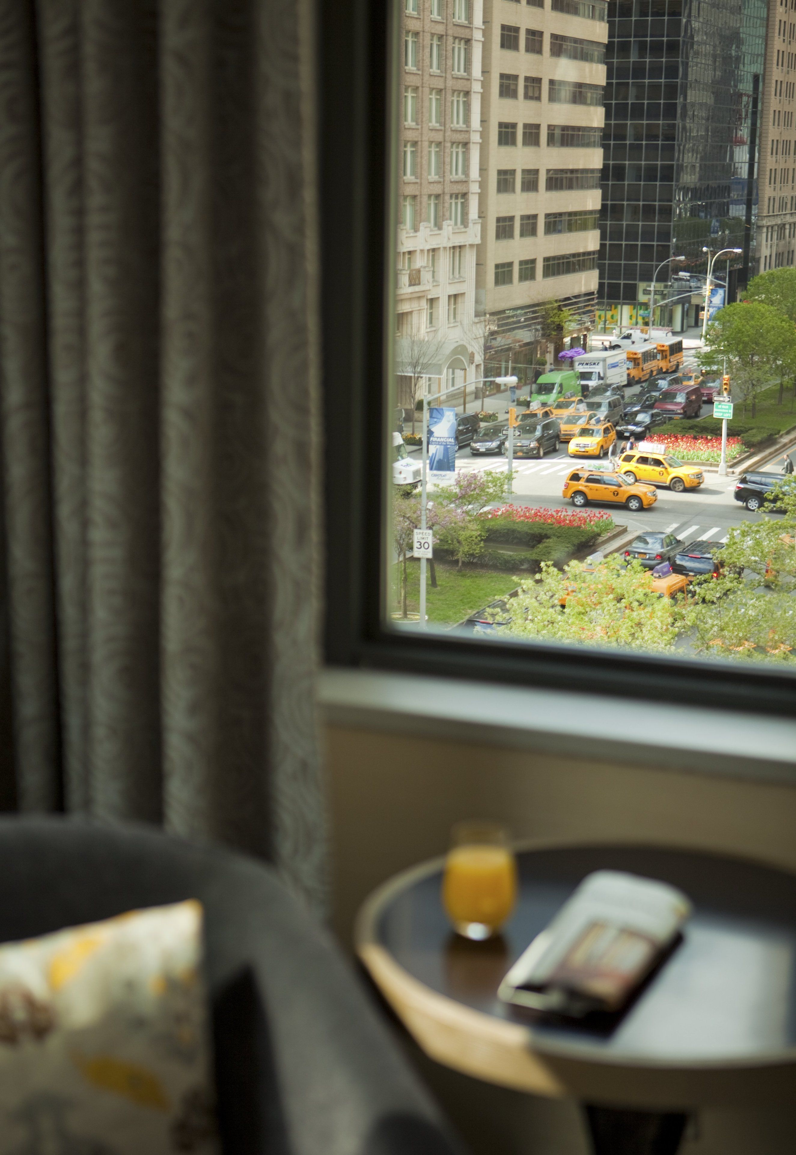 Habitación de hotel con vista a una calle concurrida a través de una ventana, una pequeña mesa con un vaso de jugo de naranja y una revista.