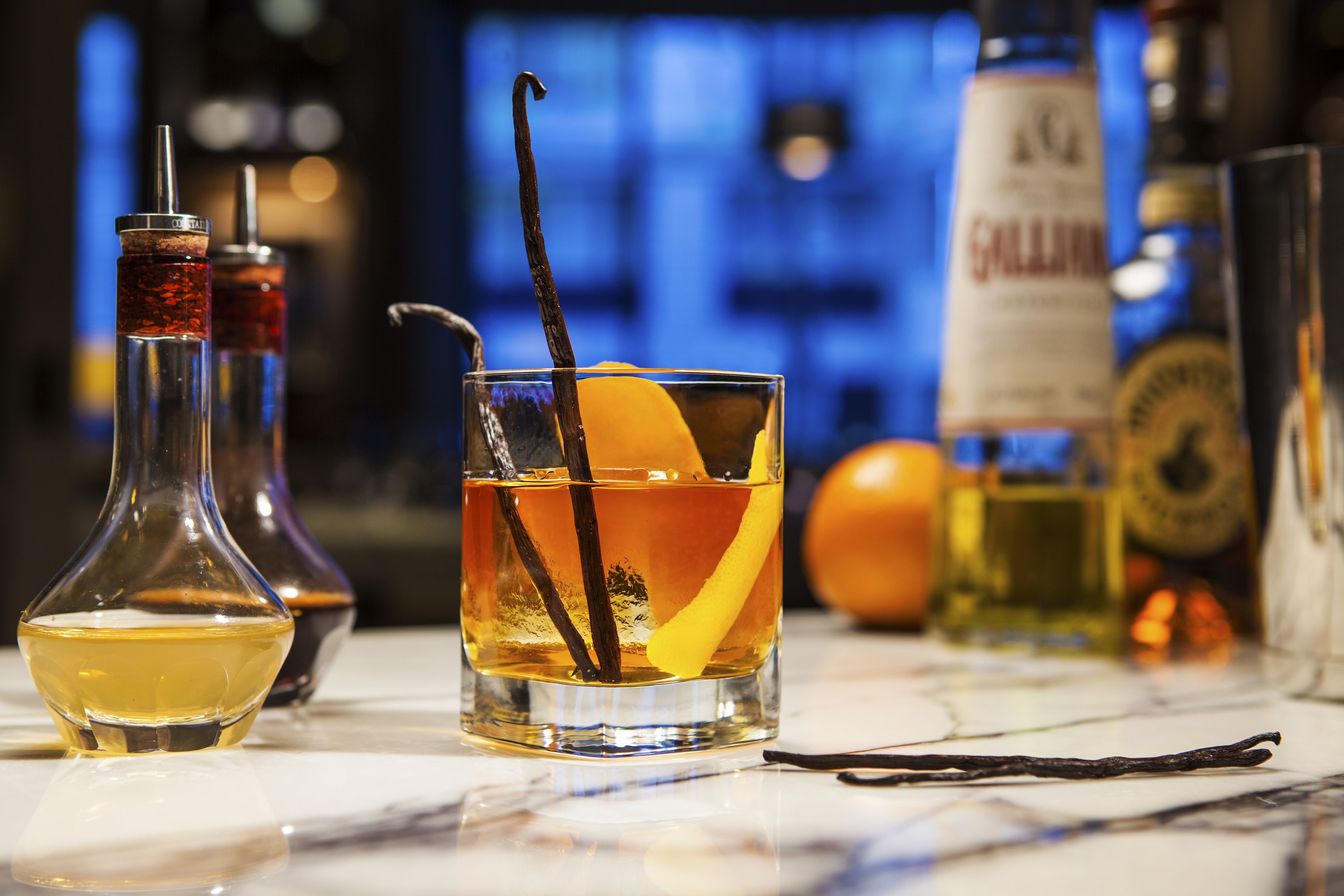 A bar setting with a glass of amber-colored drink garnished with an orange peel and a vanilla bean, accompanied by a bottle of whiskey and other bottles in the background.