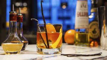 A bar setting with a glass of amber-colored drink garnished with an orange peel and a vanilla bean, accompanied by a bottle of whiskey and other bottles in the background.
