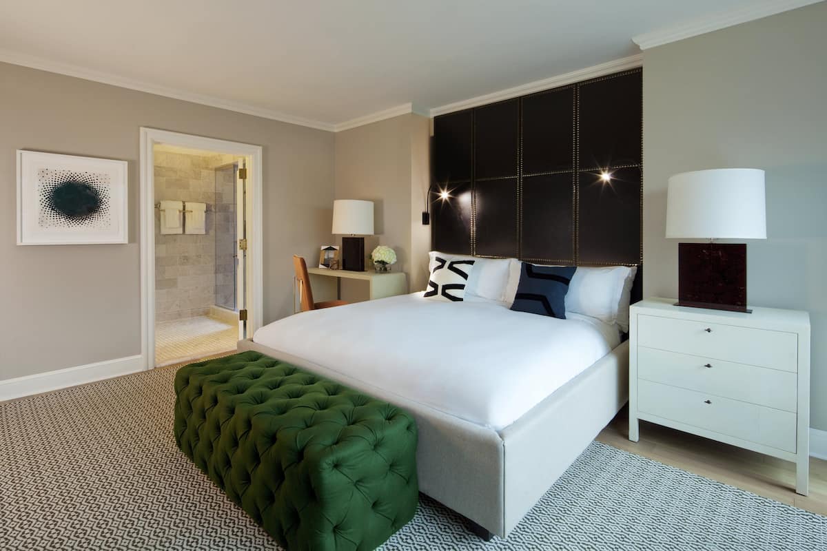 A modern hotel room with a large bed, a green tufted ottoman, a white nightstand, and a bathroom visible through an open door.