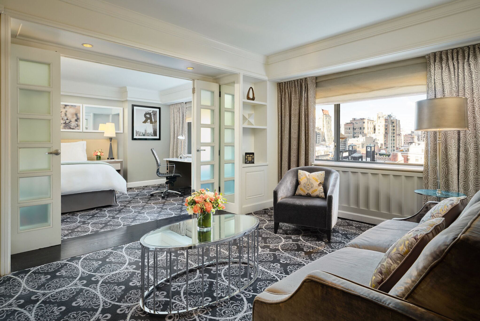 A hotel room with a bed, a sofa, a glass coffee table, a desk, and a view of the city.