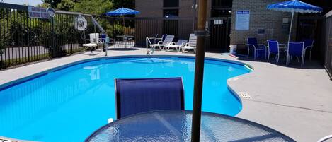 A clear blue swimming pool with lounge chairs and umbrellas, surrounded by a fence and a building with a brick facade.