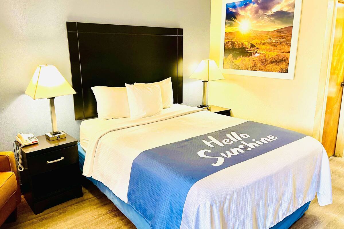 A hotel room with a bed, two bedside tables with lamps, a nightstand with a phone, a blue and white bedspread with "Hello Sunshine" text, and a landscape painting on the wall.