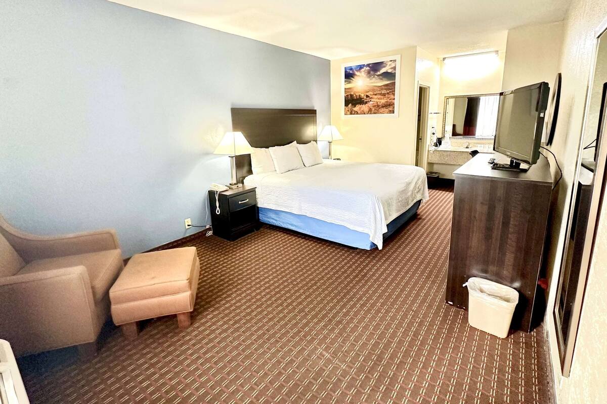 A hotel room with a bed, two bedside lamps, a chair, a desk with a computer, a TV, and a waste bin.
