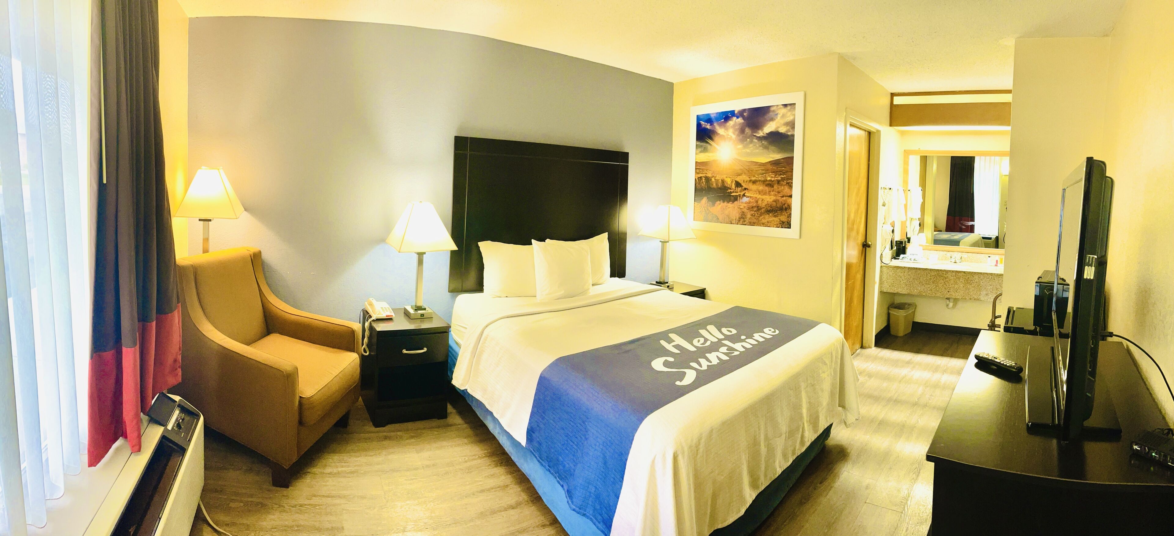 Een hotelkamer met een bed, een stoel, een bureau met een computer, een lamp en een schilderij aan de muur.