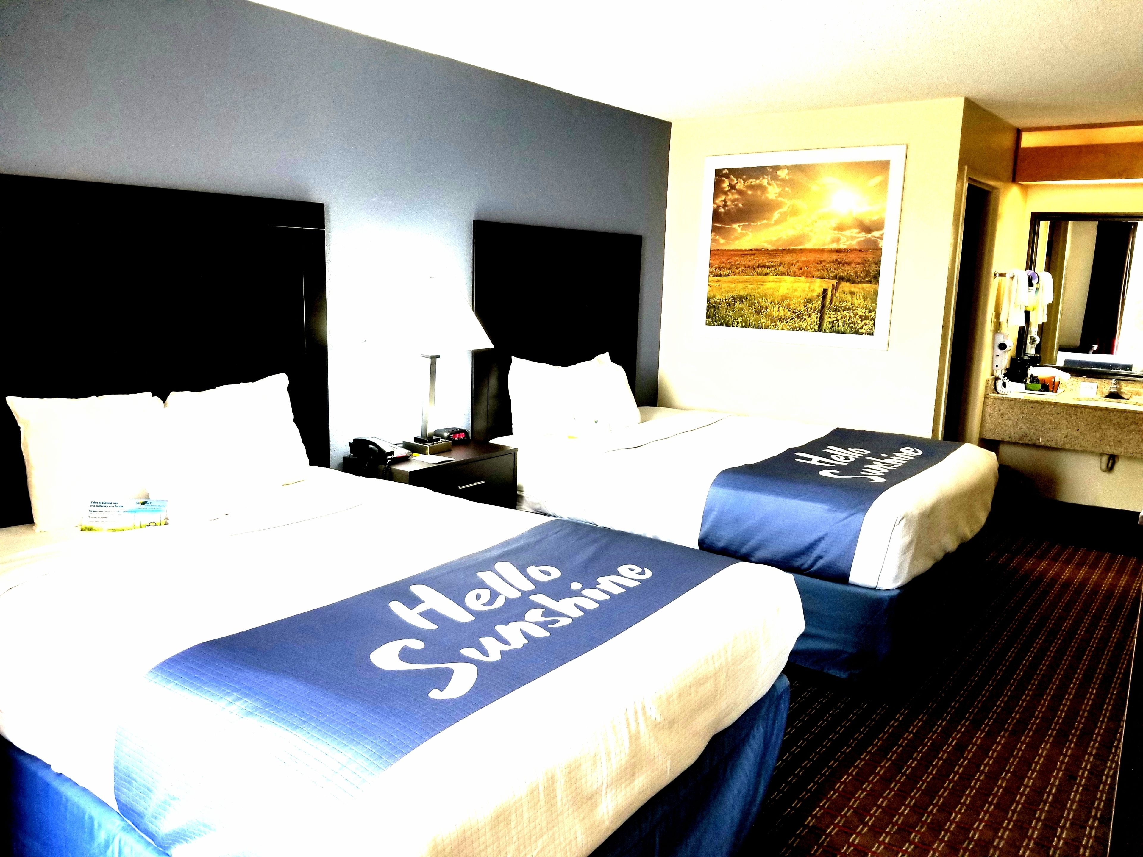 A hotel room with two beds, each with a 'Hello Sunshine' bedspread, a nightstand with a lamp, a bedside table, and a window with a view of a sunset.