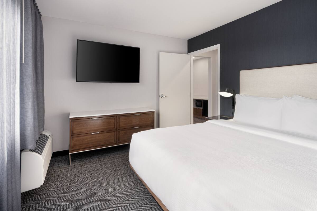 A hotel room with a bed, a flat-screen TV mounted on the wall, a wooden dresser, and a door leading to another room.