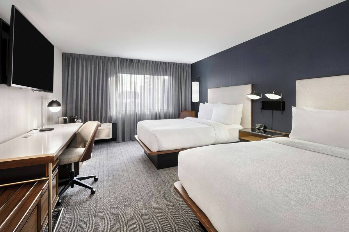 A hotel room with two beds, a desk, a chair, a flat-screen TV, and a large window with curtains.