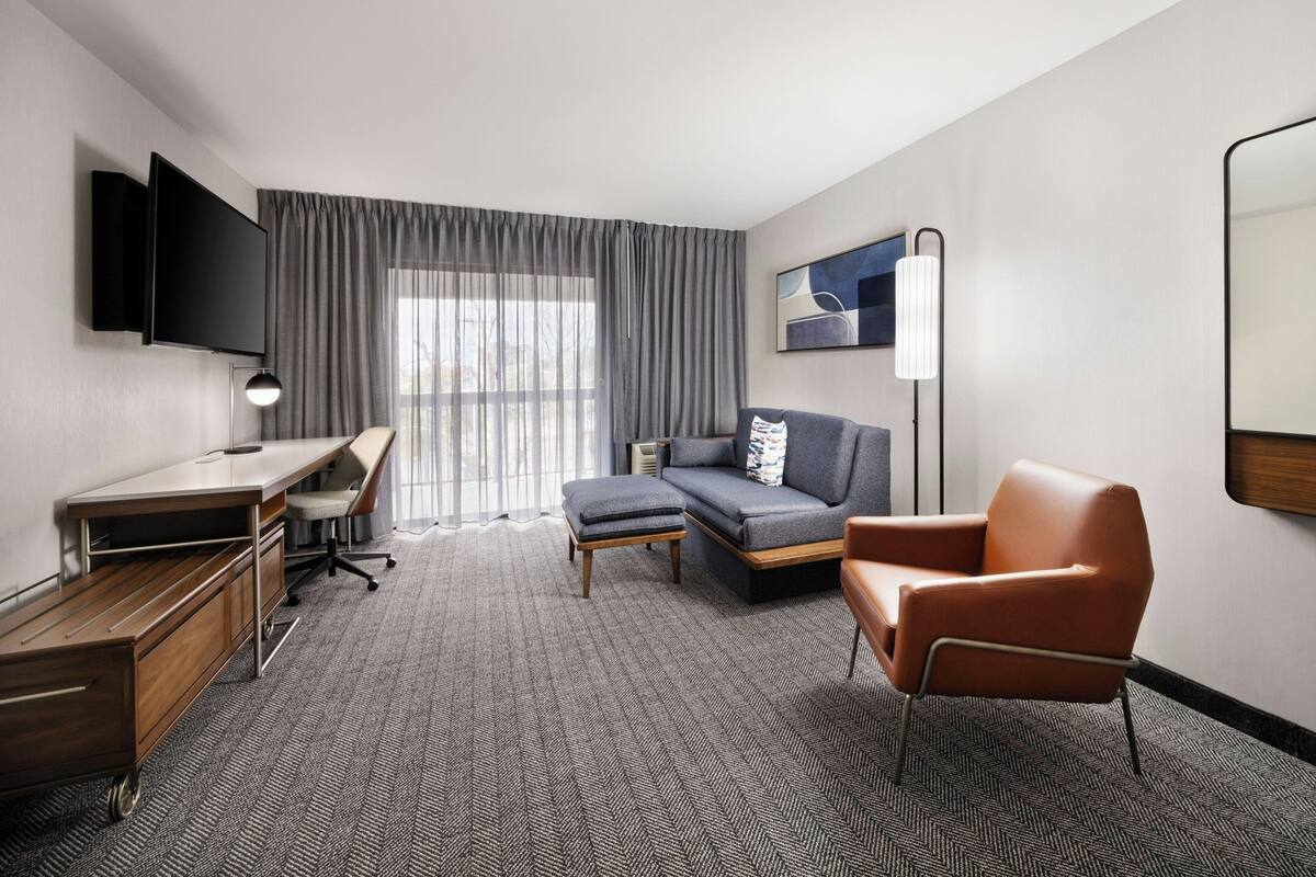 A modern hotel room with a sofa, armchair, desk, and flat-screen TV.