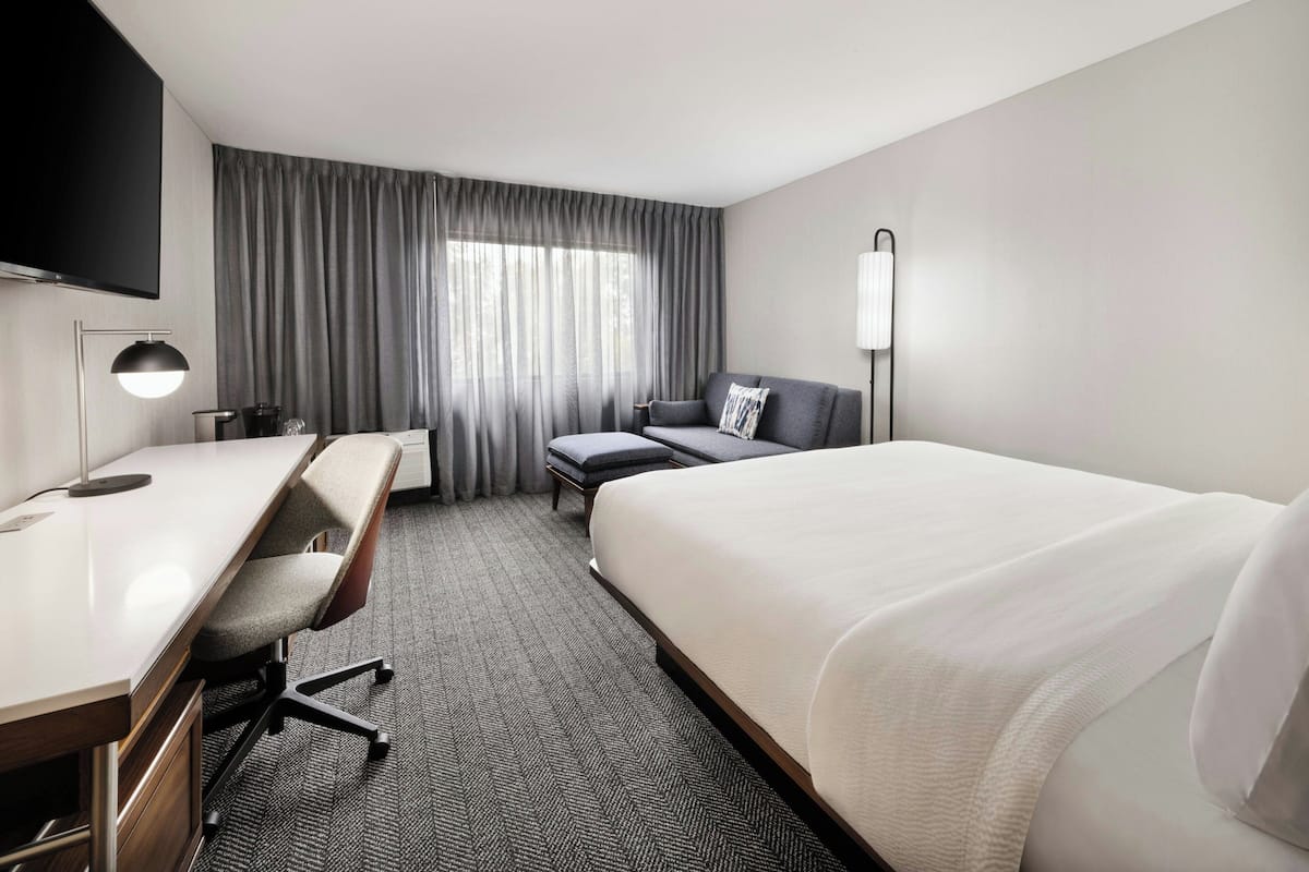 A hotel room with a bed, a desk with a chair, a TV, and a window with curtains.