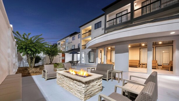 A modern outdoor patio with a stone fire pit, seating, and a multi-story building in the background.