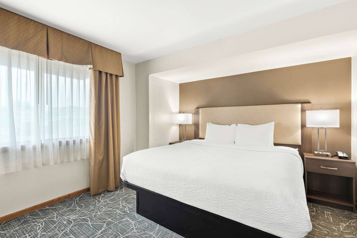 A hotel room with a large bed, two bedside tables with lamps, a headboard, and a window with curtains.