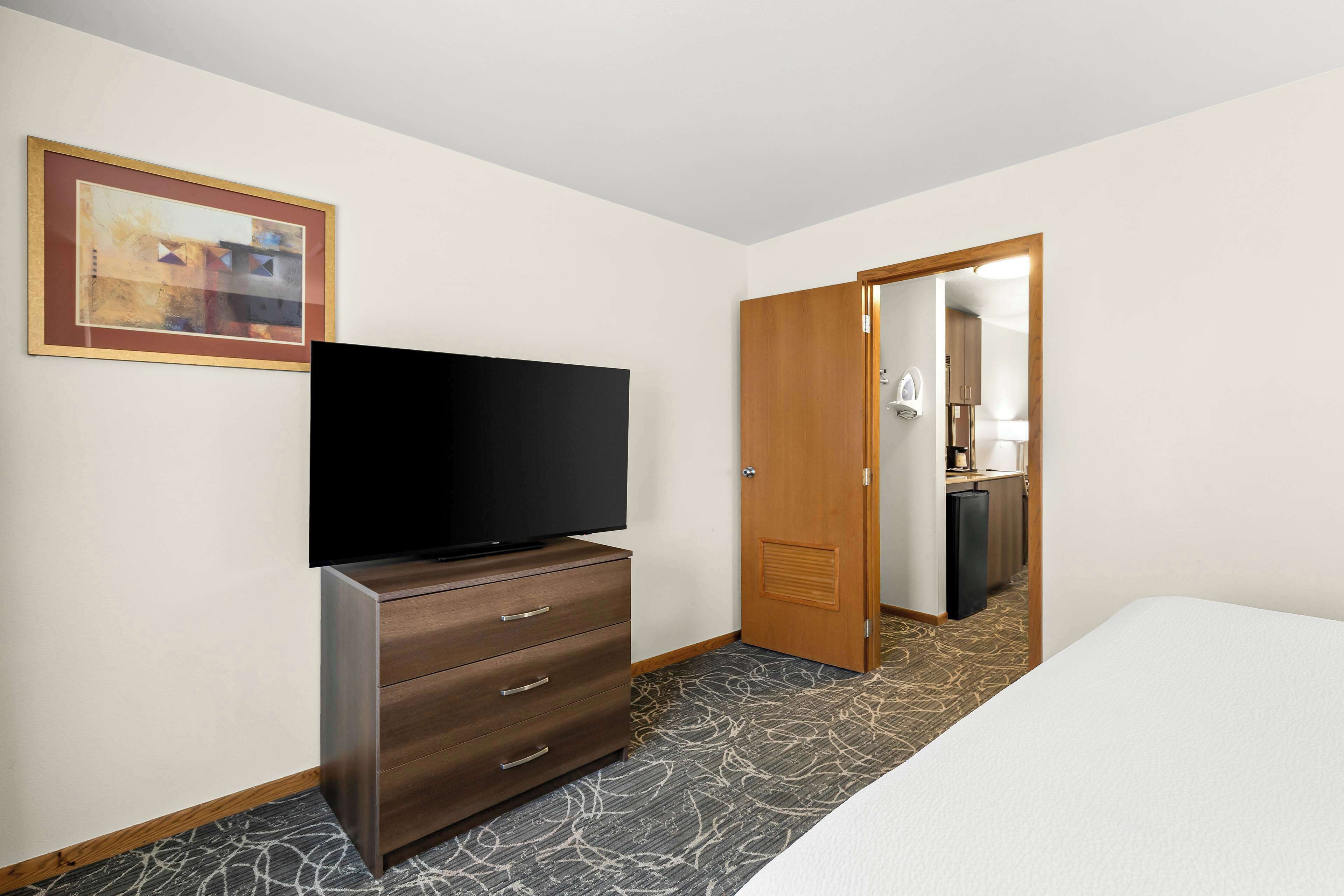 A hotel room with a flat-screen TV on a dresser, a bed, a door leading to a bathroom, and a framed picture on the wall.