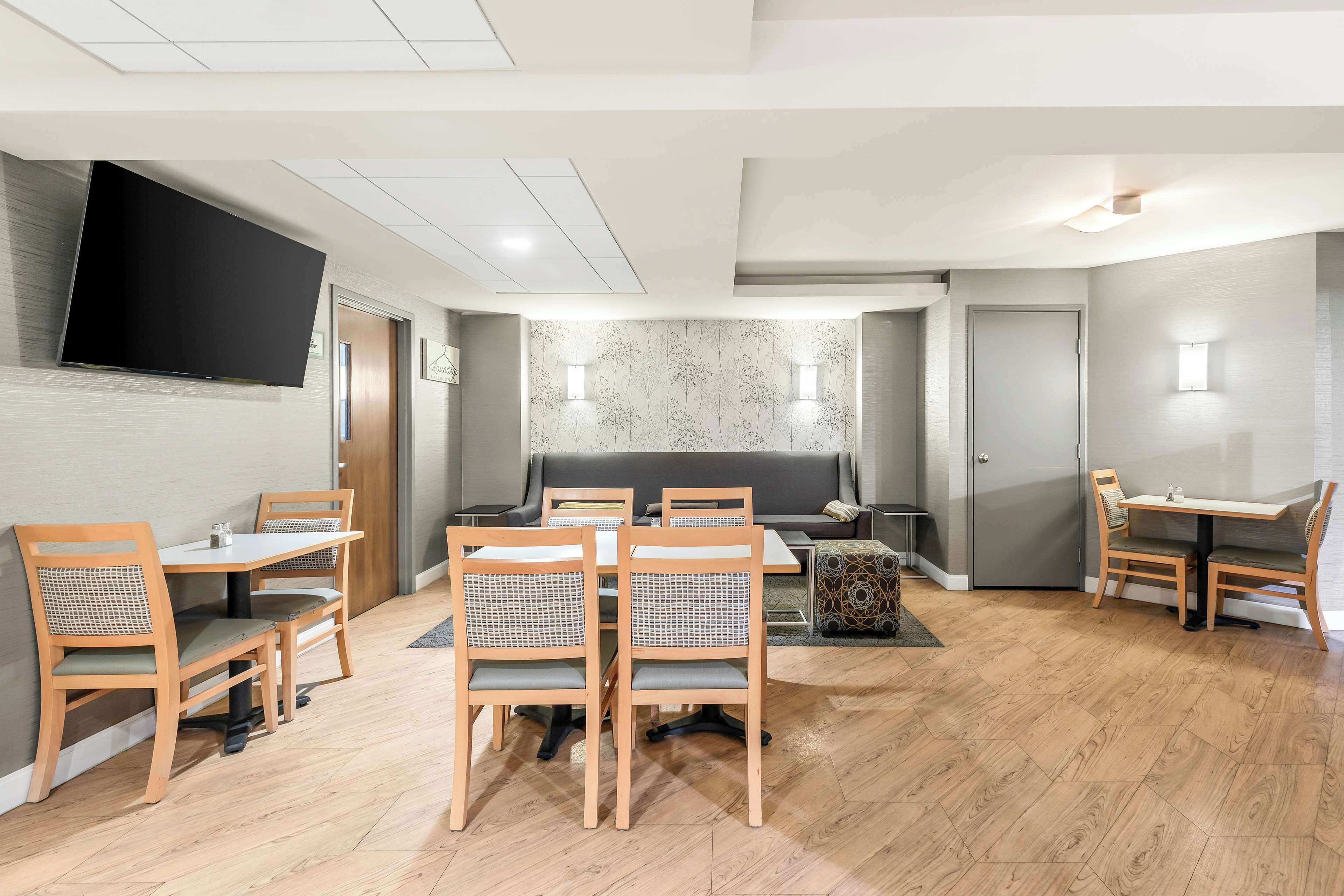 A modern dining area with wooden tables and chairs, a flat-screen TV mounted on the wall, and a patterned wall behind the seating.