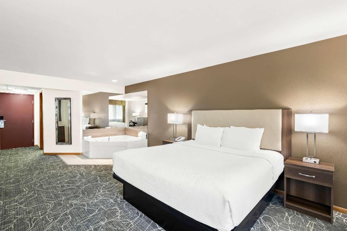 A hotel room with a large bed, two bedside tables, a nightstand, a bathroom with a mirror, and a small seating area.
