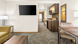 Habitación de hotel con televisión de pantalla plana montada en la pared, un sofá, una mesa de comedor y una kitchenette con armarios y un microondas.