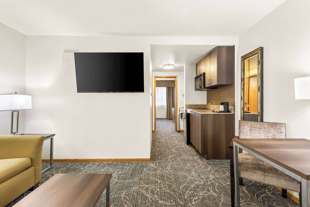 A hotel room with a flat-screen TV mounted on the wall, a sofa, a dining table, and a kitchenette area with cabinets and a microwave.