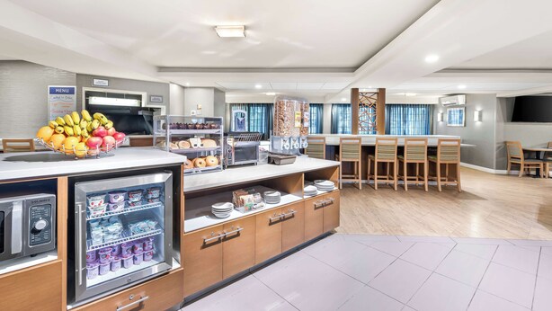 Un comedor de hotel moderno con una estación de desayuno autoservicio, un refrigerador con bebidas y un bufé con diversas opciones de comida.