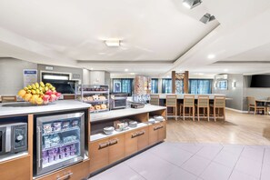 A modern hotel dining area with a self-service breakfast station, a refrigerator with beverages, and a buffet with various food items. - Best Western West Towne Suites (Madison)