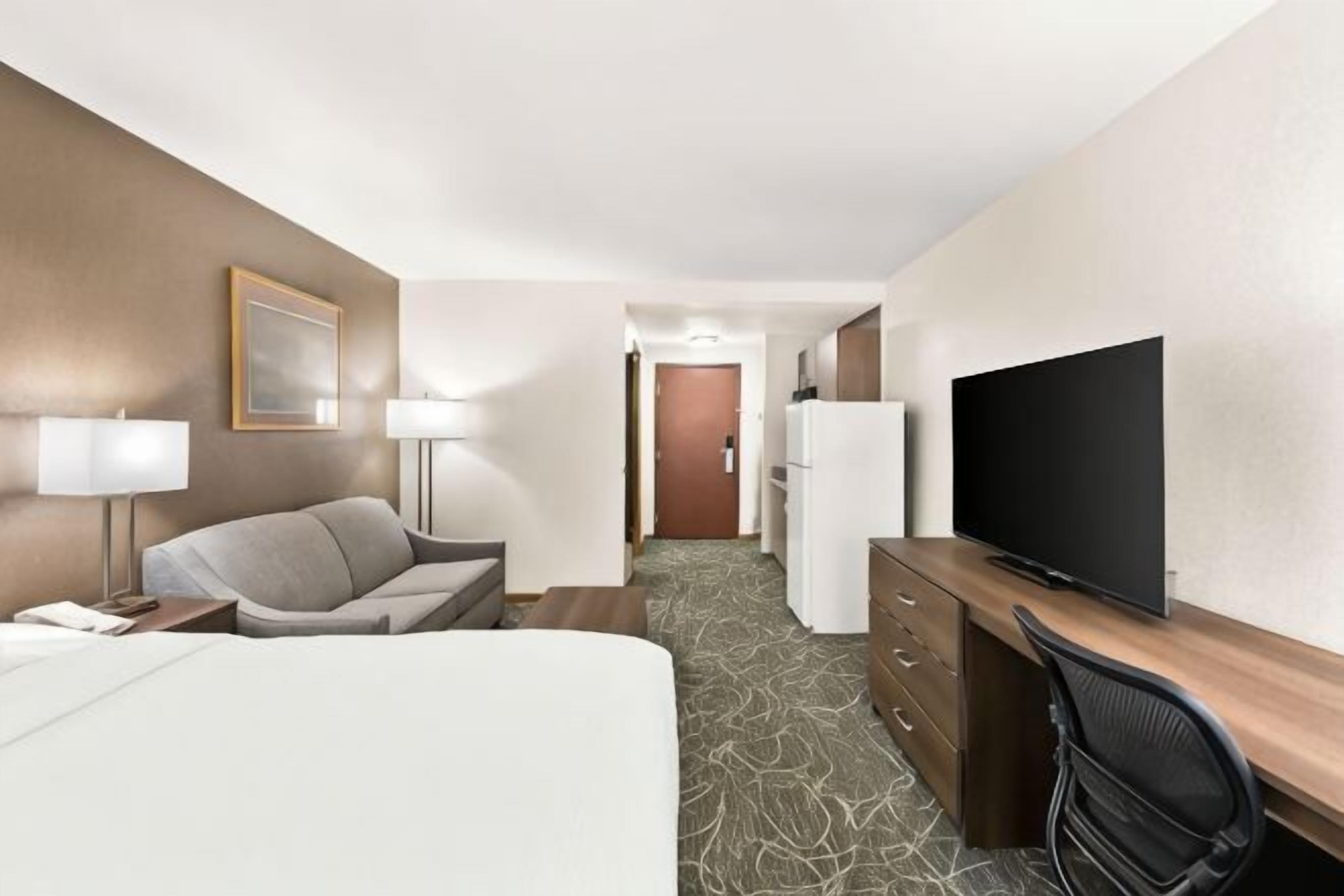 A hotel room with a bed, a desk, a television, a chair, a lamp, and a door leading to a hallway.