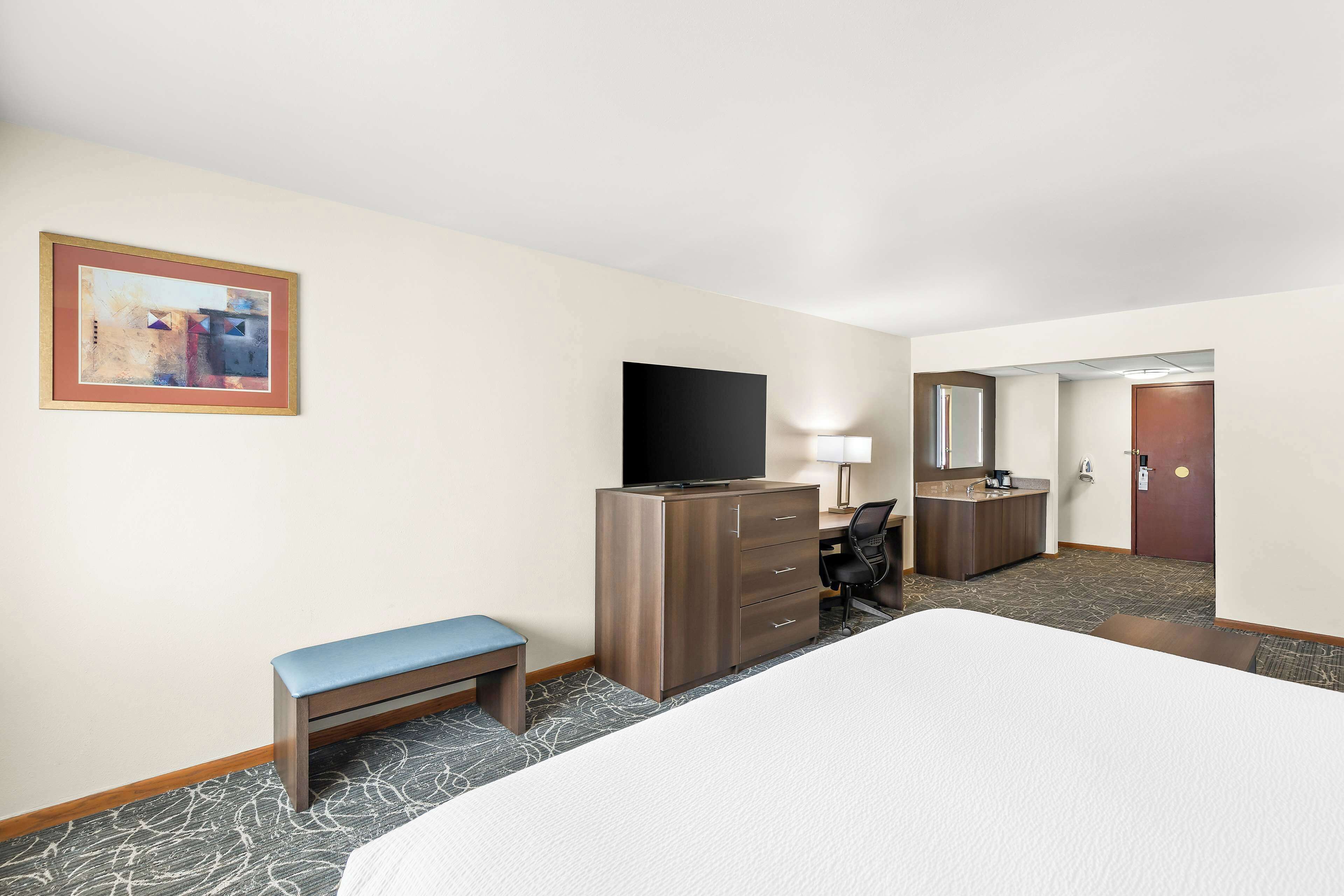 A hotel room with a large bed, a desk with a computer, a television, a bench, and a dresser.